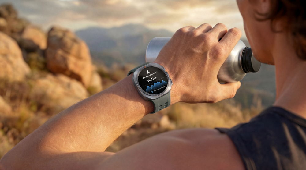 A imagem mostra uma pessoa de costas, com foco em seu pulso erguido e no Samsung Galaxy Watch Ultra2. O mostrador no relógio exibe o recurso Corrida em Trilha, indicando a distância percorrida. Na mão esquerda, a pessoa segura uma garrafa prateada, e o fundo da imagem remete a algum ambiente montanhoso.