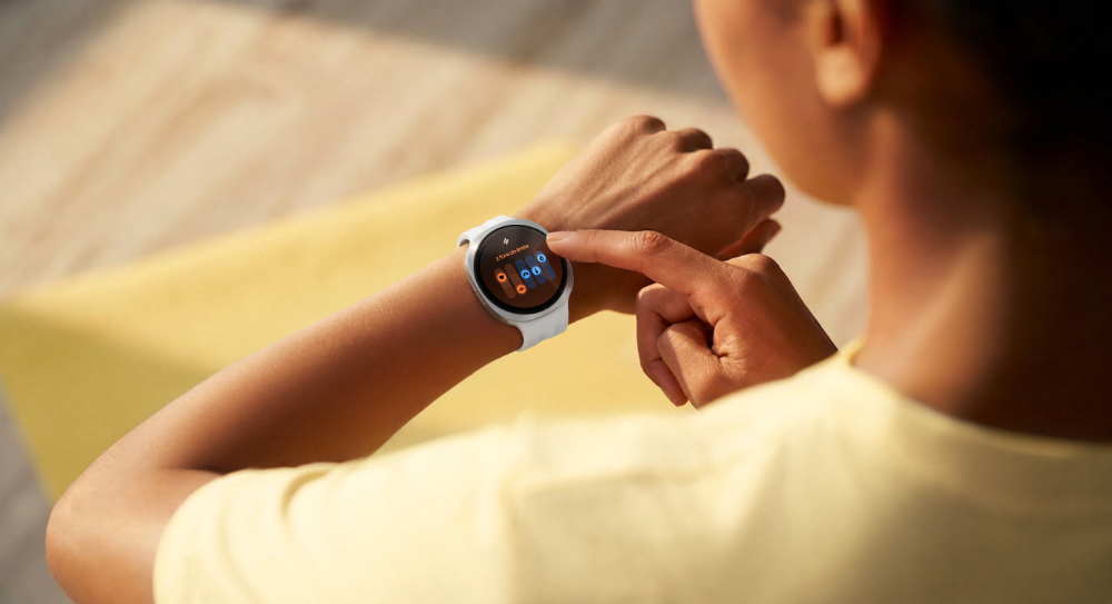 Uma pessoa aparece de costas mexendo em um Samsung Galaxy Watch9 em seu pulso esquerdo. A pessoa usa o dedo indicador da mão direita para usar a tela do relógio, que por sua vez exibe o recurso "Sinais Vitais". A pessoa usa uma camiseta amarela e está iluminada pela luz do sol.